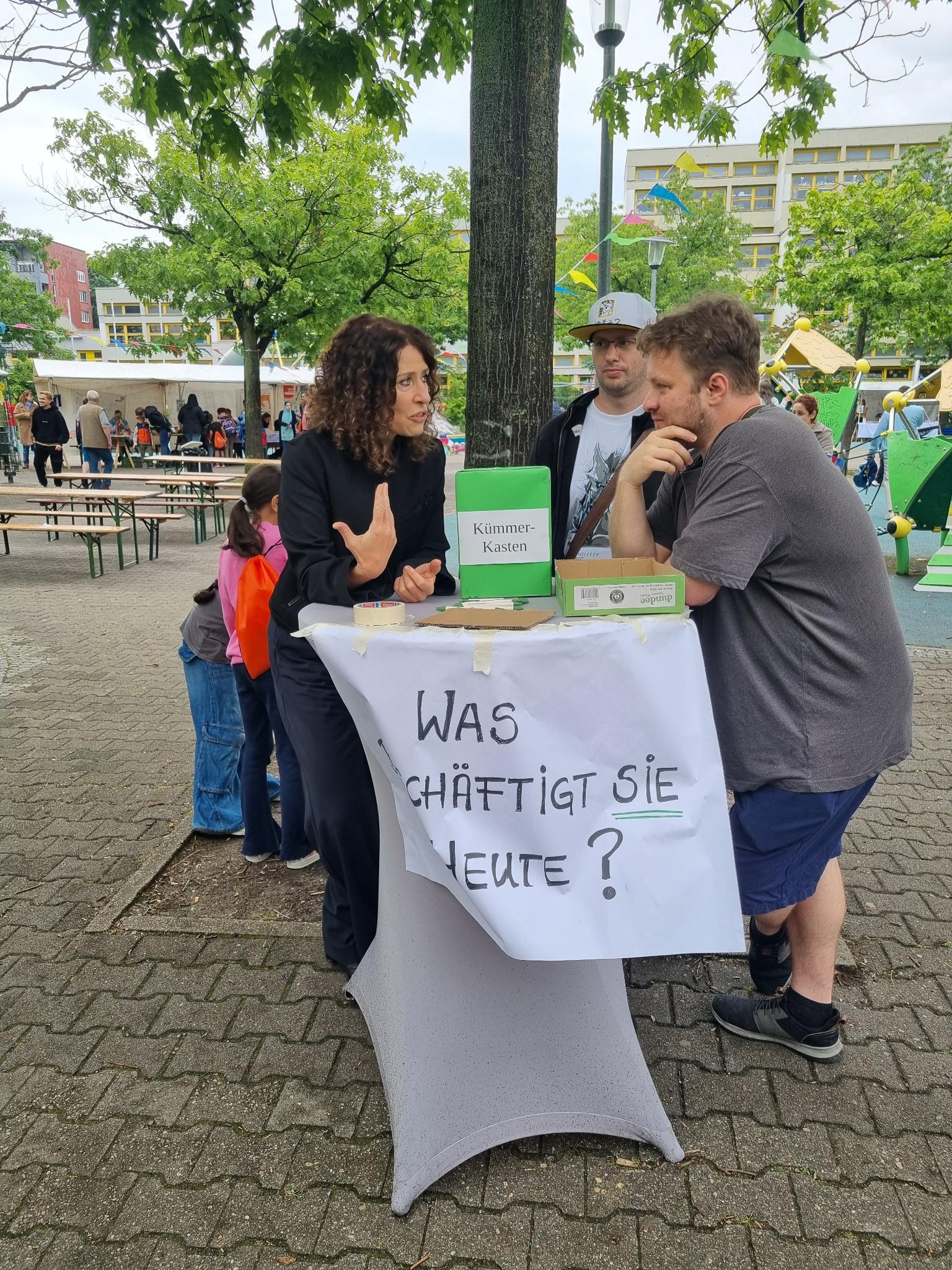 Bettina Jarasch, Nico und Johannes stehen am einem Tisch und diskutieren. Auf dem Tisch steht eine Kiste mit Aufschrift "Kümmerkasten" in den man Zettel werfen kann. Am Tisch hängt ein Plakat mit der Frage: Was beschäftigt Sie?